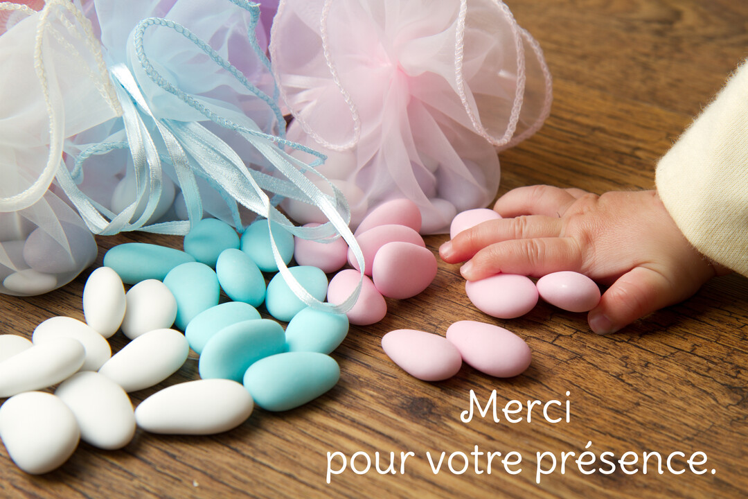 Une main de bébé touche délicatement des dragées roses, bleues et blanches étalées sur une surface en bois à côté de petits sachets en tulle.