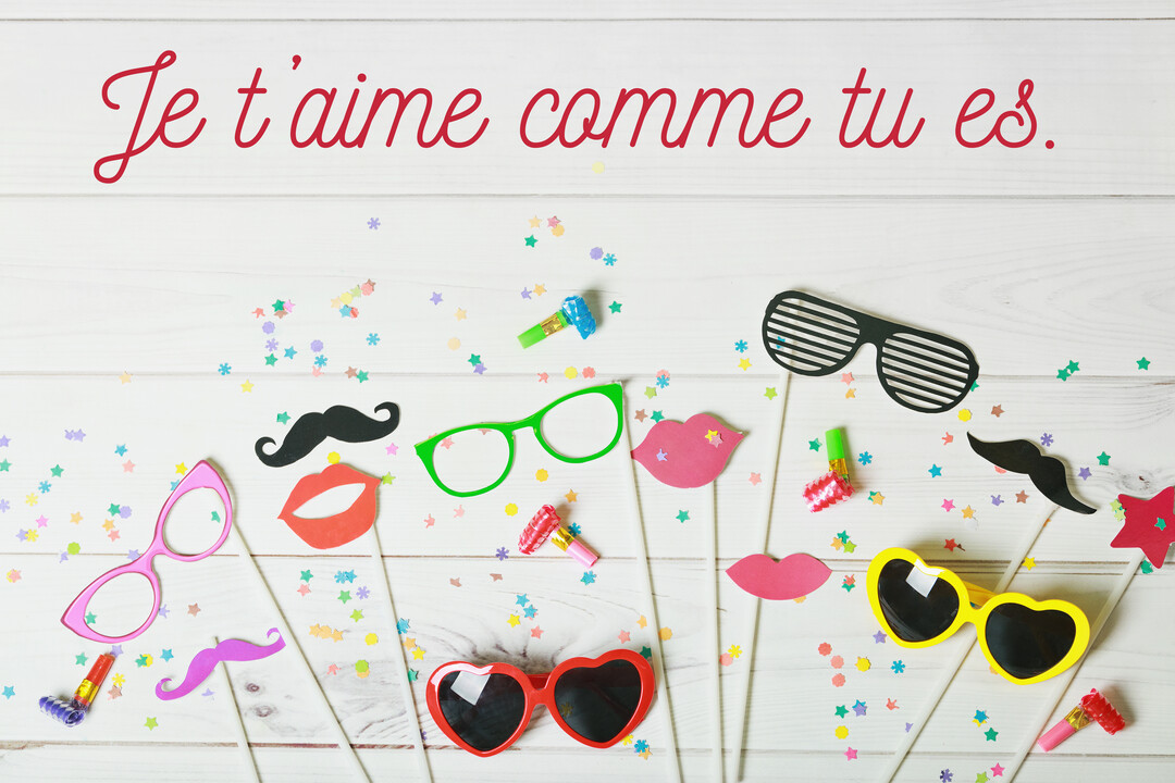 Une composition festive à plat d'accessoires de photobooth colorés sur un fond en bois blanc, avec le message d'amour manuscrit 'Je t'aime comme tu es' en rouge.